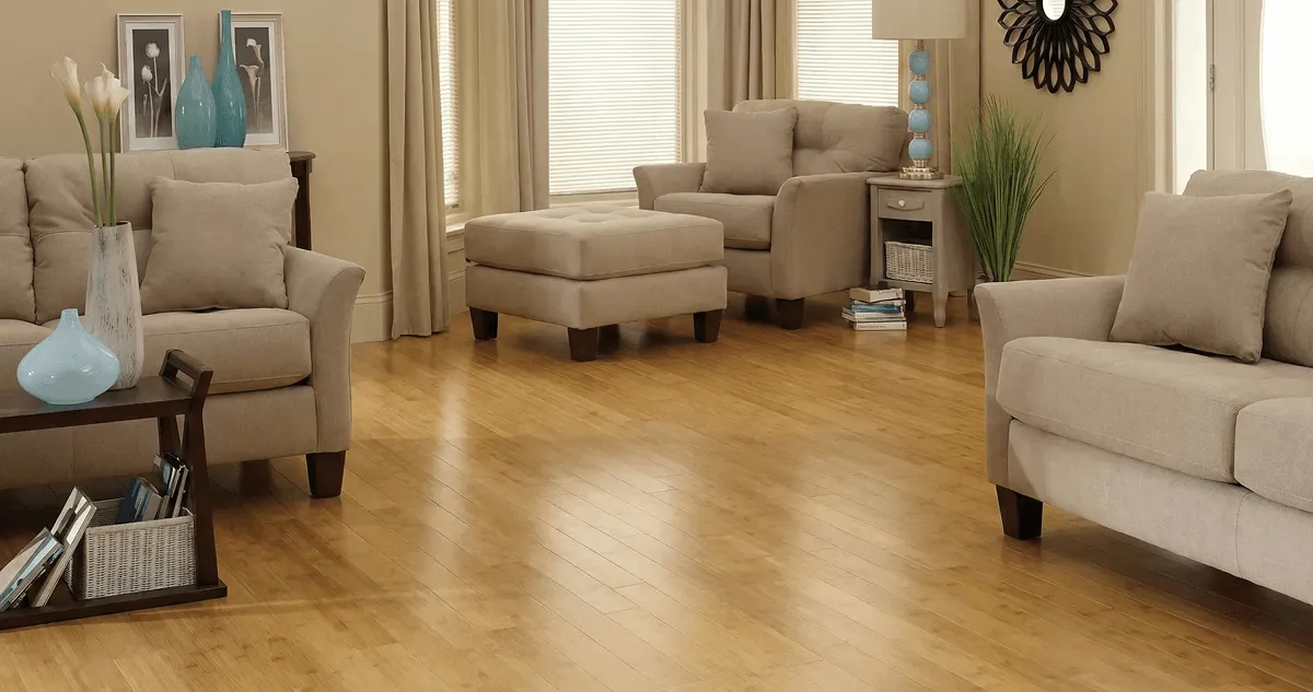 A living room with professionally installed bamboo flooring, beige sofas, a matching ottoman, a side table with a blue lamp, vases, framed pictures, and large windows with blinds and curtains in Mesa, AZ